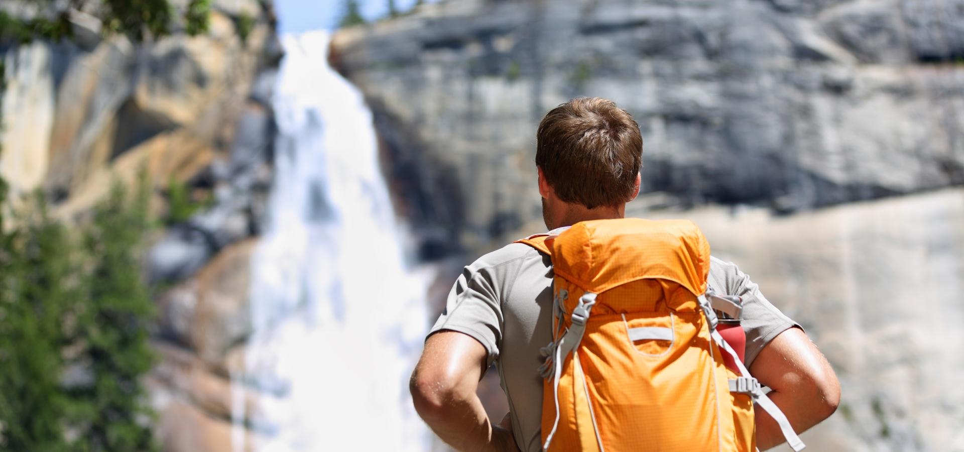 Hiker hiking with backpack looking at waterfall in Yosemite park in beautiful summer nature landscape. Portrait of male adult back standing outdoor.